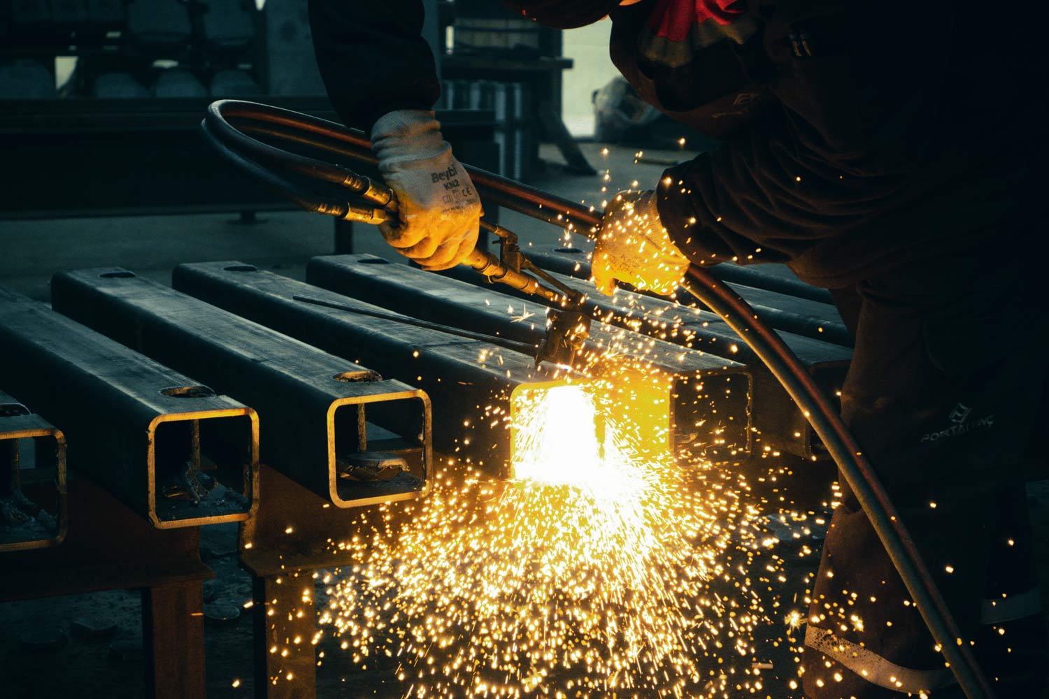 A fabricator cutting steel box sections with a torch, sparks flying across the bench